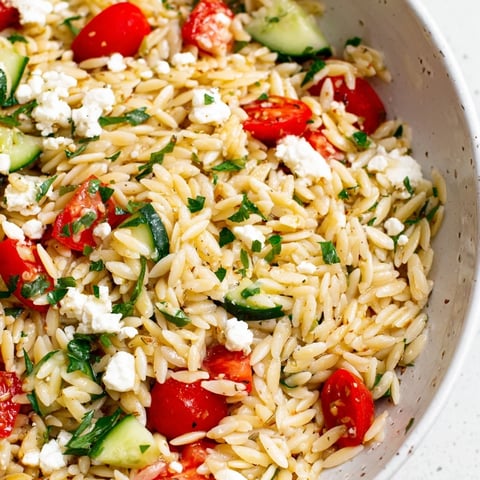 A close-up of Greek Orzo Salad with feta crumbles, halved cherry tomatoes, diced cucumber, and Kalamata olives on a rustic wooden table.  
