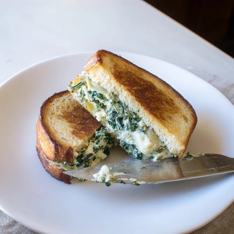 Two halves of a golden, cheesy Spinach Artichoke Grilled Cheese rest on a plate, ready for a comforting lunch.  